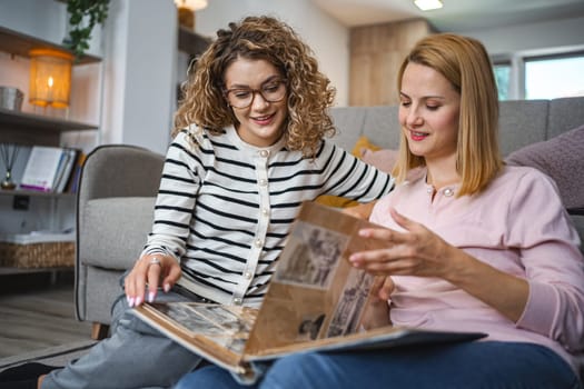 Two female friends are sitting on the floor in the living room, smiling and looking at and flipping through an old photo album.