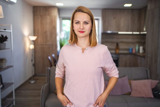 A woman is standing next to a couch in a living room. She is looking around the room, seemingly contemplating her surroundings. The room is cozy and well-furnished, with various decorations and a soft rug on the floor.