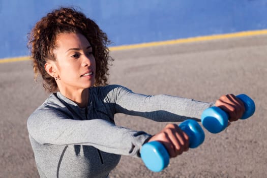 female athlete in sportswear working out with dumbbells during training on asphalt road, concept of urban sport and healthy lifestyle, copy space for text