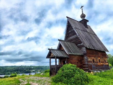 A serene wooden church stands on a hillside, framed by lush greenery and a river below, with dramatic clouds enhancing the atmosphere.