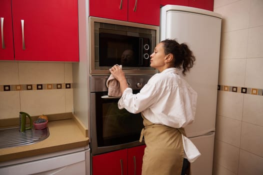 A woman in an apron cleans her kitchen oven with a cloth. The kitchen has bright red cabinets and a sleek design. The scene captures domestic chores and housekeeping.