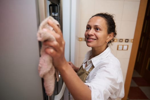 A cheerful woman wearing an apron cleans the kitchen, wiping down surfaces with a cloth. Her smile conveys satisfaction and cleanliness. The modern kitchen setting adds a homely feel.