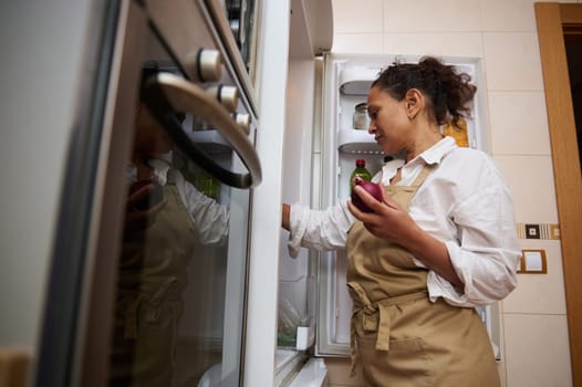 A woman in an apron stands in a kitchen, holding an onion, while looking inside an open fridge. She appears focused and ready to prepare a meal.