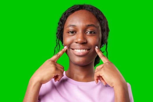 Smiling young woman in a pink shirt pointing to her cheek against a bright green background in studio