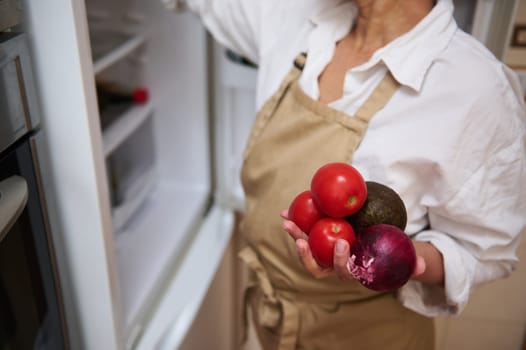 A woman stands near an open refrigerator, holding fresh vegetables in her hand. She wears a white shirt and a beige apron, suggesting a focus on cooking or meal preparation.