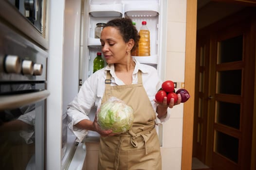 A woman in a kitchen apron selects fresh vegetables from a refrigerator, preparing for a meal. The image conveys a homely and healthy lifestyle, emphasizing nutrition and culinary preparation.
