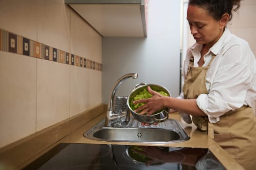 A woman rinses fresh lettuce in a modern kitchen. She wears an apron while preparing ingredients, reflecting a calming domestic scene. Perfect for illustrating healthy home cooking or lifestyle.
