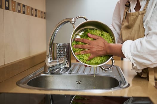 A home chef wearing an apron washes fresh green lettuce in a colander over a modern kitchen sink. The hands-on preparation highlights fresh ingredients essential for healthy home cooking.