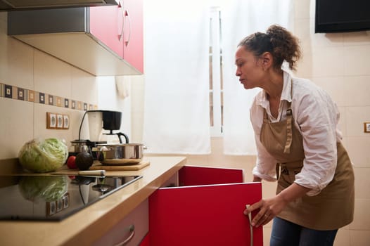 A woman wearing an apron is seen organizing her kitchen. Fresh vegetables, a coffee maker, and a pot line the counter, depicting a warm, homely kitchen atmosphere.