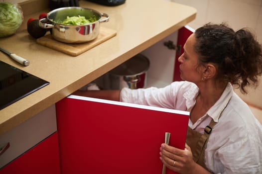A woman in a kitchen prepares a meal, opening a red cabinet while making a fresh avocado salad. The setup includes various kitchen tools and a promising healthy cuisine vibe.