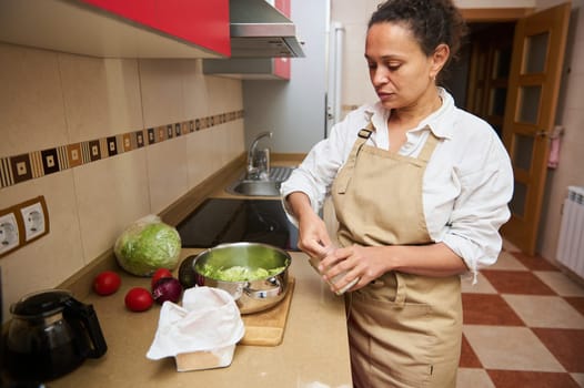 A woman in an apron is preparing fresh vegetables on a kitchen counter. The kitchen has modern fittings and natural lighting. She appears focused on her cooking task.