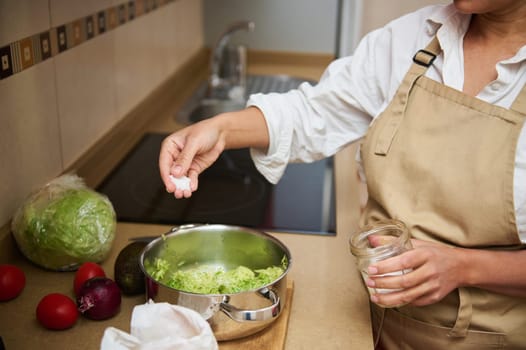 A woman in an apron seasons fresh guacamole in a modern kitchen, surrounded by avocados, tomatoes, and lettuce.