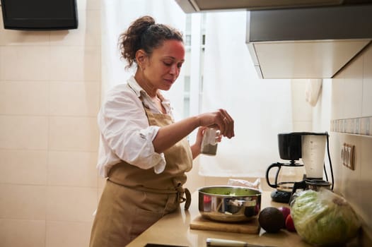 A woman in an apron adds seasoning to a fresh salad in a modern kitchen. The setting is cozy and bright, with a focus on healthy cooking and home lifestyle.