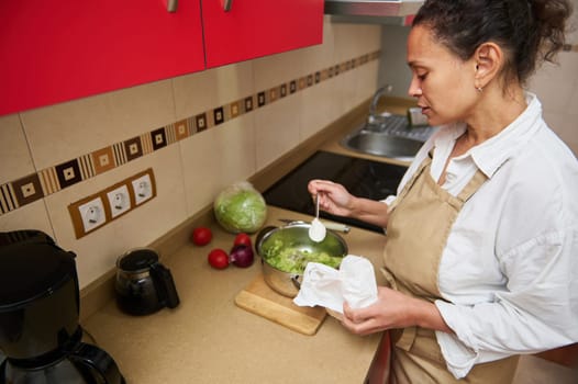 A woman in an apron is mixing a fresh salad in a modern kitchen setting, adding seasoning with care. Ingredients such as lettuce, onions, and tomatoes are visible on the countertop.