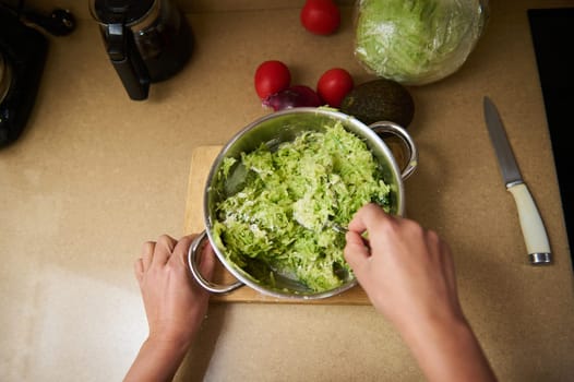 Hands preparing a fresh green salad in a bowl, surrounded by various vegetables on a kitchen countertop. A knife and cutting board are also visible, indicating a cooking process.