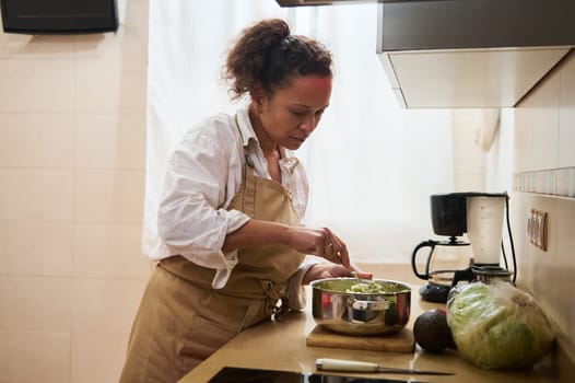 A woman wearing an apron mixes fresh ingredients in a stainless steel bowl, creating a salad in a brightly lit kitchen, embodying health, home cooking, and culinary passion.