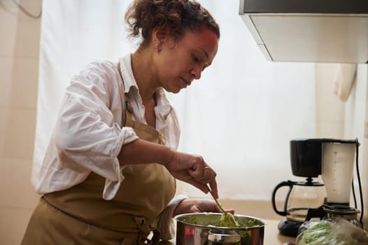Woman cooking in a kitchen, carefully preparing a fresh meal. Dressed in an apron, she stirs ingredients in a pot, emphasizing home cooking and culinary passion.