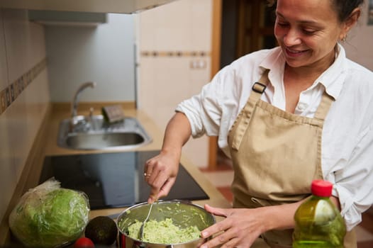 Woman smiling while making guacamole in her kitchen. Ingredients include avocado and vegetables. Culinary creativity and healthy eating concept.