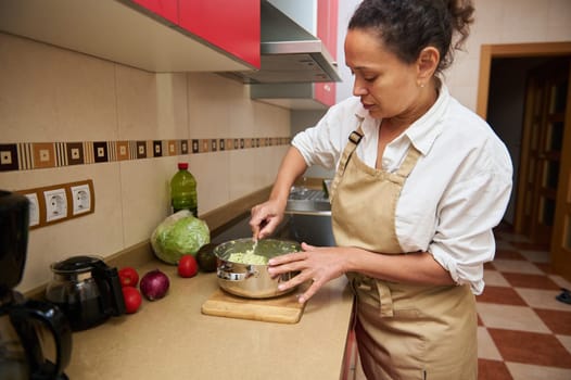 A woman in an apron mixes vegetables in a pot, preparing a fresh salad in a modern kitchen setting.