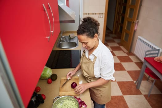 A woman wearing an apron slices an onion in a modern kitchen. Surrounded by fresh vegetables like avocados and tomatoes, she prepares a salad with focus and creativity.