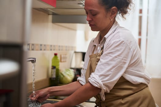 A woman wearing an apron washes her hands at the kitchen sink, ensuring cleanliness and hygiene. The running water and kitchen setting emphasize a focus on healthy home practices.