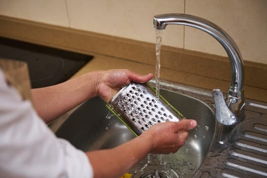 Close-up of hands cleaning a metal grater under running tap water. The process of washing kitchen utensils after use, promoting cleanliness and hygiene.
