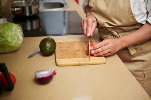 A person in an apron slices a red bell pepper on a wooden cutting board. Fresh ingredients like avocado, lettuce, and onion are nearby, emphasizing a focus on healthy cooking.