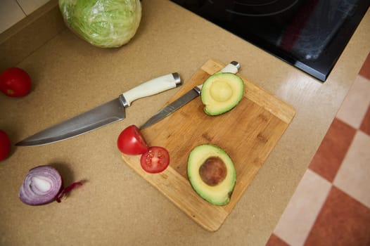Sliced avocado and tomato on a cutting board, with a knife on a kitchen countertop. Nearby are a whole lettuce and half a red onion, creating an inviting, fresh cooking scene.