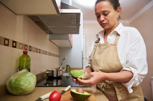 A woman in a kitchen carefully peeling an avocado, surrounded by fresh ingredients. She wears an apron, suggesting a focus on healthy cooking and meal preparation.