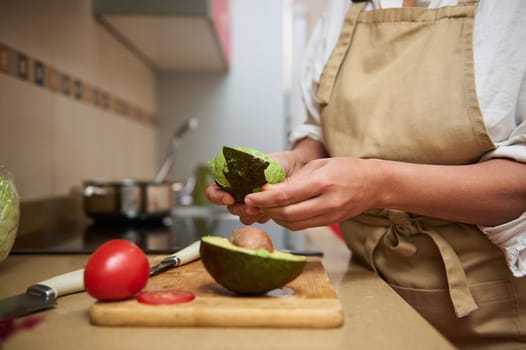 A person wearing an apron is peeling a ripe avocado on a wooden cutting board in a kitchen setting. Ingredients like tomato and lettuce are visible, suggesting a healthy meal preparation.