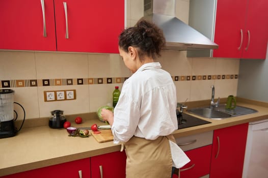 A woman in a kitchen prepares fresh vegetables on a countertop. The kitchen features modern red cabinets and appliances, creating a vibrant atmosphere. Cooking and healthy living concept.