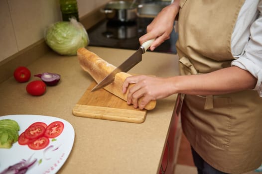 A woman slices a baguette on a wooden cutting board, preparing a fresh sandwich with vegetables in a kitchen setting. Ingredients, including tomatoes and onions, are visible.