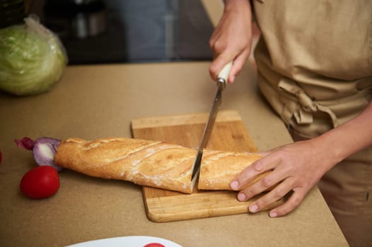 A person wearing an apron is slicing a fresh baguette on a wooden cutting board in a kitchen. The scene contains ingredients like onion and tomato, suggesting a wholesome meal preparation.