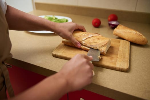 A close-up of a person slicing a fresh baguette on a wooden cutting board, surrounded by fresh vegetables like tomatoes and avocado, in a cozy kitchen setting.