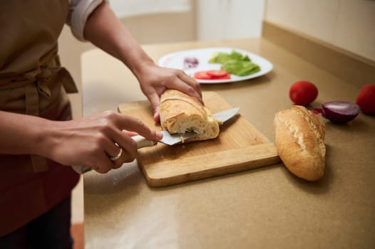A person slices a baguette on a wooden cutting board in a warm kitchen setting, with fresh vegetables nearby.