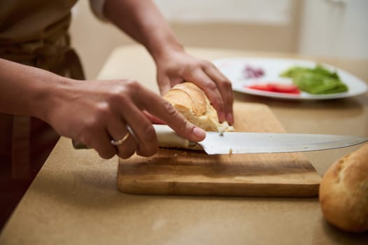 A person skillfully slices a crusty loaf of bread on a wooden cutting board, with a plate of fresh vegetables visible in the background.