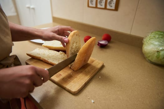 Close-up of hands slicing crusty bread on a wooden board. Fresh vegetables like tomatoes and lettuce are in the background, suggesting preparation for a meal. Warm kitchen ambiance.