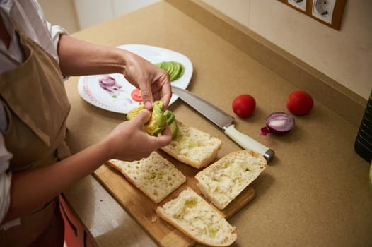 A person prepares fresh avocado sandwiches in a cozy kitchen, slicing ingredients and arranging them on bread. The scene evokes a sense of healthy, homemade meal preparation.