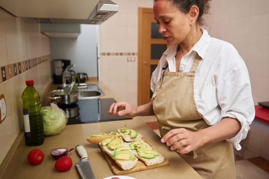 A woman in an apron makes avocado sandwiches in a home kitchen. Fresh ingredients like tomatoes and lettuce are on the counter, embodying a healthy cooking vibe.