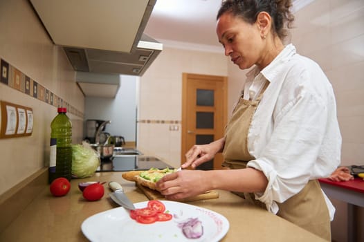 A woman in an apron prepares a fresh sandwich in a modern kitchen, surrounded by vegetables. She is focused on creating a healthy meal, embodying the concept of home cooking and nutrition.