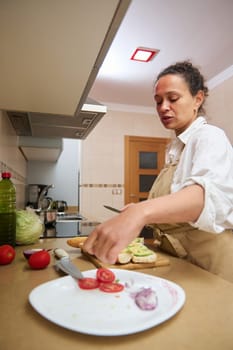 A woman in an apron prepares fresh vegetables in a modern kitchen, slicing tomatoes and placing them on bread. The atmosphere is casual and focused, highlighting home cooking and culinary skills.