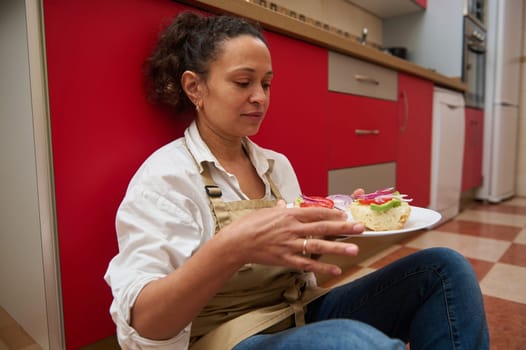 A woman relaxing on the kitchen floor with a homemade sandwich on a plate. She looks content, enjoying a moment of quiet in a cozy kitchen. The setting is casual and inviting.