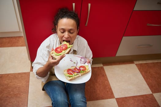 A person seated on the kitchen floor indulges in avocado toast topped with fresh tomatoes and onions, capturing a relaxed and carefree moment.