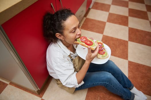A woman sits on a kitchen floor savoring a fresh sandwich with avocado and tomato slices, capturing a moment of relaxation and enjoyment. The casual setting emphasizes comfort and contentment.