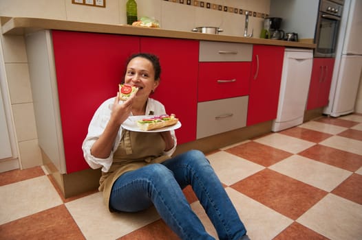 A cheerful woman sits on a kitchen floor, enjoying a homemade sandwich. She is relaxed with a joyful expression, showcasing a casual and content moment in a home environment.