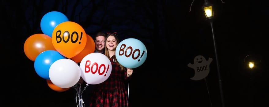 A cheerful young couple stands in a dark setting, holding colorful balloons that read BOO! The festive atmosphere captures the spirit of Halloween festivities and joy.