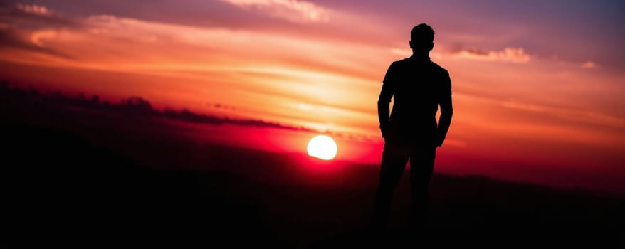 A lone individual stands on a hilltop, gazing at the vibrant sunset. The sky is filled with hues of orange and purple as the sun dips towards the horizon, creating a breathtaking view.