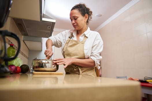 Focused woman in apron cooking in a modern kitchen, stirring ingredients in a pot. Fresh vegetables are seen on the counter.