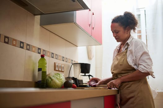 A woman is focused on preparing a nutritious meal in a well-lit kitchen, wearing an apron. Fresh vegetables and cooking ingredients are visible in the peaceful, homey space.