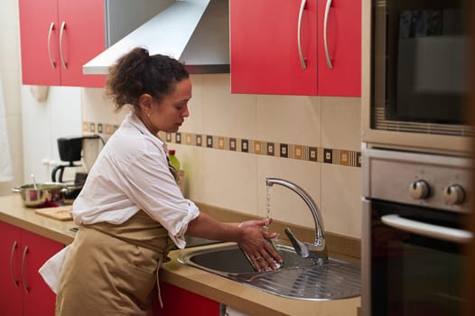 A woman cleans her hands under running water in a modern kitchen with red cabinets. The scene emphasizes cleanliness and hygiene, highlighting the significance of washing hands in daily routines.